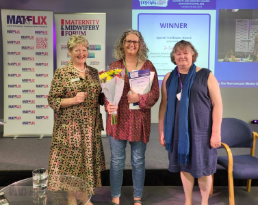 Colour photo of three women - Jo Dagustun in the centre - at a maternity conference. Jo is holding a bunch of flowers and some papers